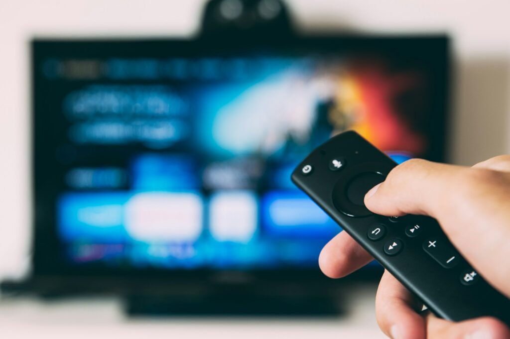 Person holding a TV remote. A TV with a streaming app is visible in the background.